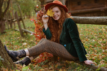Happy smiling redhead woman hides her eye with yellow autumn leaf. Model wearing trendy orange hat,...
