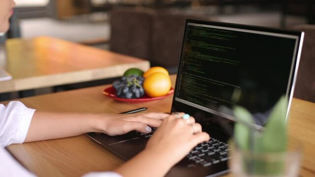 Back view over the shoulder shot of developer programmer female with laptop. Program code and script data on the screen. Young freelancer woman working on project in cafe.