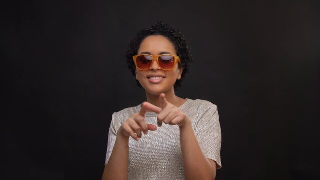 leisure and people concept - happy african american woman in sunglasses dancing and drawing heart in air with fingers over black background