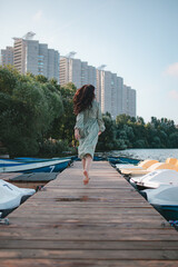 a girl in a light dress barefoot runs along the pier near the water, a boat, a beautiful girl runs in a dress