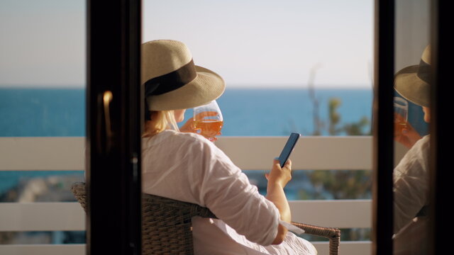 Woman with wine glass using cell when relaxing at the balcony overlooking sea