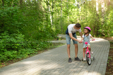 Fototapeta premium Happy father teaching his little daughter to ride a bicycle. Child learning to ride a bike. Family activities at summer.