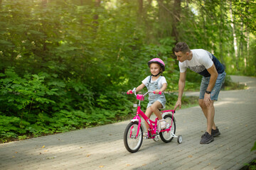 Happy father teaching his little daughter to ride a bicycle. Child learning to ride a bike. Family activities at summer.