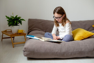 Obraz premium a cute little girl in glasses, jeans and a white turtleneck is reading a book, doing homework, preparing for exams sitting on the bed. home education. space for text. High quality photo