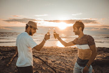 Cheerful young people spending time together on the beach and drinking beer and toasting at the beach on vacation in twilight summer sunset. Friendship concept