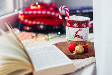 A mug of coffee with a deer,  snowflakes with marshmallows, a candy cane, an open book on the windowsill on a background of warm sweaters and garlands. Cozy winter Christmas evening. New Year's Eve