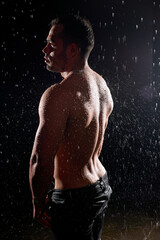 Fototapeta na wymiar Side view on sexy muscular sportive man with strong body, showing muscular back standing isolated against black background, in rain, water drops on body. Caucasian european male model