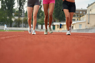 Cropped shot of legs of three diverse female runners in sportswear running together on track field at the stadium