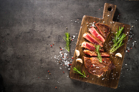 Grilled Beef steak at wooden cutting board with spices. Top view with copy space at black background.