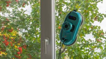 A window cleaner robot washes the window glass against the background of a garden with rowan trees...