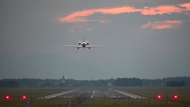 Small private jet airplane landing at Ljubljana Airport, Slovenia. Telephoto perspective of luxury aircraft approaching and descending. Colorful summer sunset. Static shot, real time