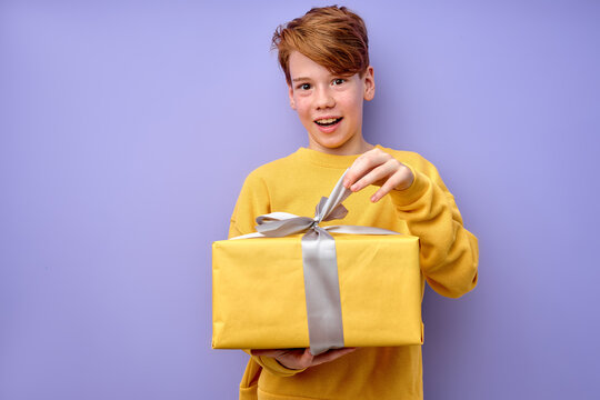 Amazed Curious Adorable Little Boy Unpacking Present Box With Funny Astonished Expression, Impatient Caucasian Child In Yellow Wear Unboxing Birthday Surprise. Studio Shot Purple Background.