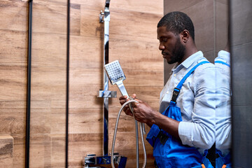 Afro american repairman handyman in blue overalls holding shower handle in hands while repairing...