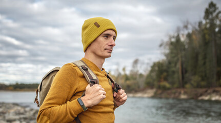 Man traveler in a yellow hat and sweater with a backpack is walking along a fast river with a stony shore