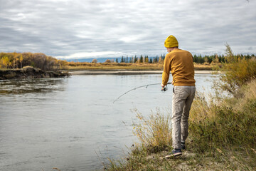 Fototapeta premium Man in a yellow sweater and hat is fishing on the river bank in the autumn forest with a spinning rod in his hands