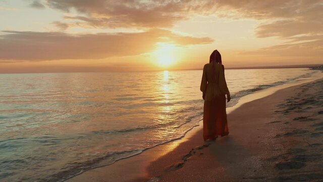 woman walking barefoot by beach at golden sunset leaving footprints in sand. Pretty woman walks at seaside surf. Splashes of water and foam. 4K 