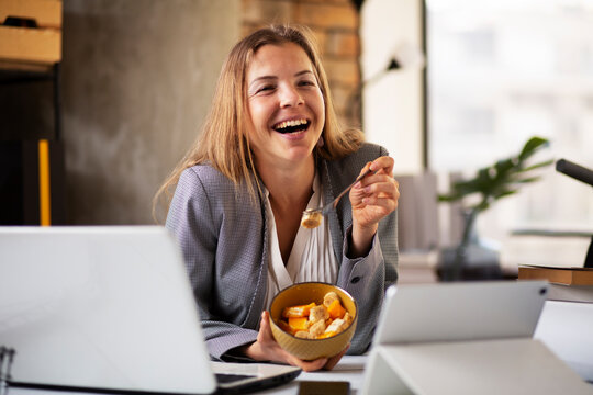 Businesswoman In Office Having Healthy Snack. Young Woman Eating Fruit While Having A Video Call