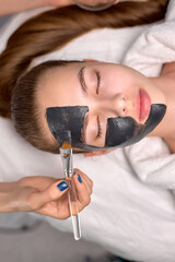 Top View On Cropped Professional Cosmetologist applying black mask on fresh face of beautiful woman for carbon peel, female is lying on bed with eyes closed. Skin care, wellbeing concept. Copy space