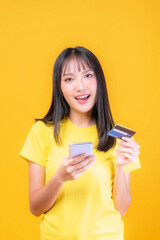 Naklejka premium beautiful Asian young woman cute girl with bangs hair style in yellow shirt holding credit card and smartphone ,mobile phone uses a online banking for pay online shopping isolated on yellow background
