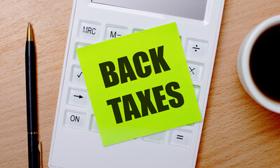 On a wooden table there is coffee in a white cup, a pen and a white calculator with a green sticker with the text BACK TAXES. Business concept