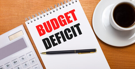 On a wooden table there is a white calculator, a white cup of coffee, a pen and a white notebook with the text BUDGET DEFICIT. Business concept
