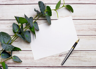 On a light wooden table, there is a eucalyptus branch, a fountain pen and a sheet of paper with a...