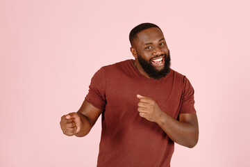 Positive bearded African-American guy actor in brown t-shirt dances standing on light pink...