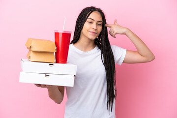 Teenager girl with braids holding pizzas and burgers over isolated pink background having doubts and thinking