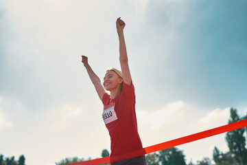 Low angle view of excited young female athlete with arms raised reaching the finish line at track field during marathon outdoors