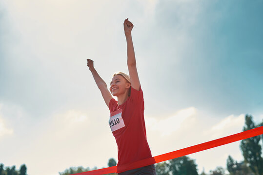 Low angle view of excited young female athlete with arms raised reaching the finish line at track field during marathon outdoors