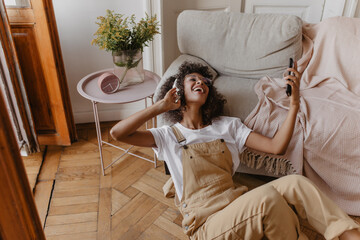 Obraz premium adorable young African woman reclining on floor in cozy light room. girl in white T-shirt and overalls listens to her favorite songs through headphones on her head, holding phone in her hands.