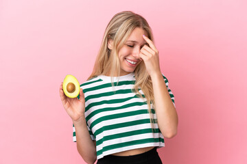 Young caucasian woman holding an avocado isolated on pink background laughing