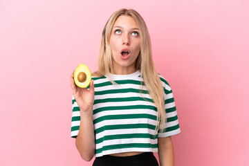 Young caucasian woman holding an avocado isolated on pink background looking up and with surprised expression