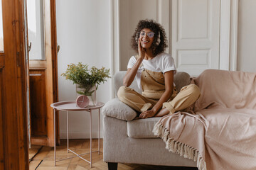 Obraz premium stunningly sweet girl sits cross-legged on white sofa, huddled in light room. swarthy girl in headphones, glasses is dressed in white T-shirt and jumpsuit on top.