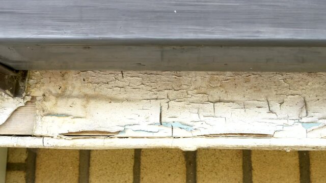Closeup POV elevated outside dolly shot at house roof level, of peeling white gloss paint on a wooden fascia board, beneath rain guttering.