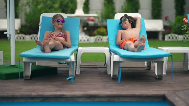 Positive laughing Caucasian boy and girl lying on sunbeds on wooden pool deck sunbathing. Wide shot portrait of cheerful friends enjoying summer vacations on sunny day outdoors on resort