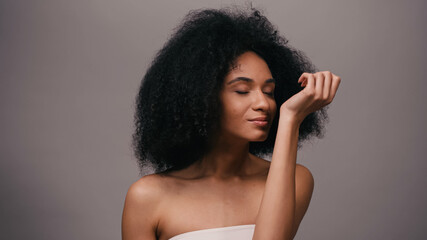 curly young african american woman enjoying odor of perfume isolated on grey