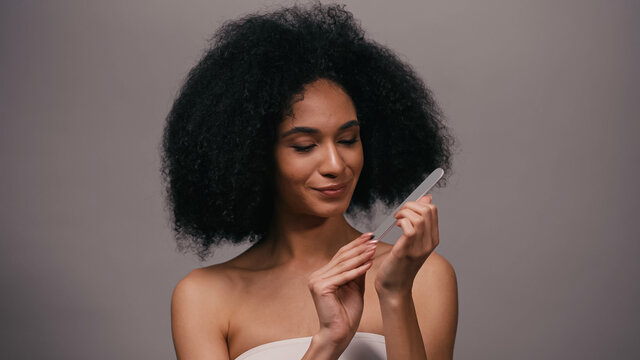 pleased young african american woman using nail file isolated on grey