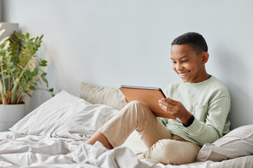 Full length portrait of smiling African-American teenager using tablet while sitting on bed in cozy...
