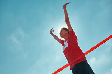 Low angle view of cheerful young female runner with arms raised reaching the finish line at track...