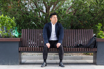 Obraz premium Asian businessman performing breathing exercises trying to calm stress, sitting on a bench during a lunch break in a business suit