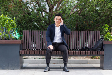 Fototapeta premium Asian businessman tired after work, sitting on a bench with fatigue and headache, has stress and panic attacks