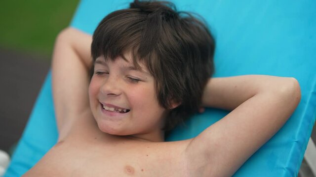 Close-up of satisfied little boy lying on blue sunbed turning smiling looking at camera. Portrait of cheerful Caucasian relaxed kid enjoying summer vacations at resort on sunny day outdoors
