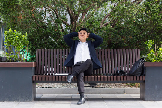 Happy asian office worker, businessman sitting and resting on bench in city park outdoors. Business man employee a formal suit break rest and relax from the outside. Downtown street, urban background