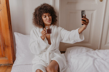 in early morning, charming beauty makes selfie to her loved one while in bed. young African woman in warm snow-white robe sits on floor with cup of fragrant latte.