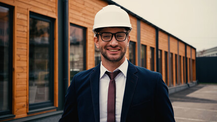 Fototapeta premium Smiling engineer in white hardhat looking at camera near building outdoors