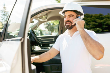 Man engineer builder wearing a white hard hat, shirt in front of his pickup using cellphone and...