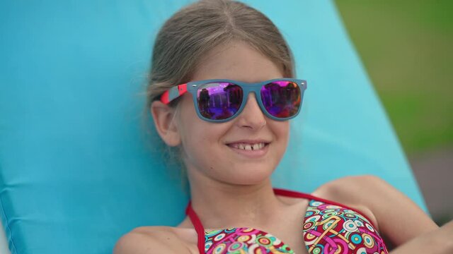 Close-up portrait of happy Caucasian girl in sunglasses sending air kiss in slow motion smiling lying on sunbed outdoors. Charming cute child posing sunbathing at summer resort on sunny day