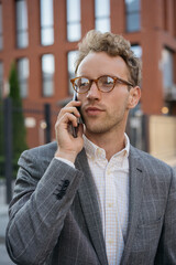 Handsome pensive  businessman wearing stylish eyeglasses talking on mobile phone standing on the street looking away