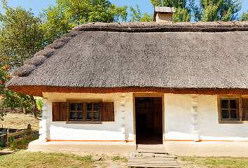 Fragment of the facade of an old traditional Ukrainian rural house with a thatched roof against the backdrop of a summer garden and blue sky.